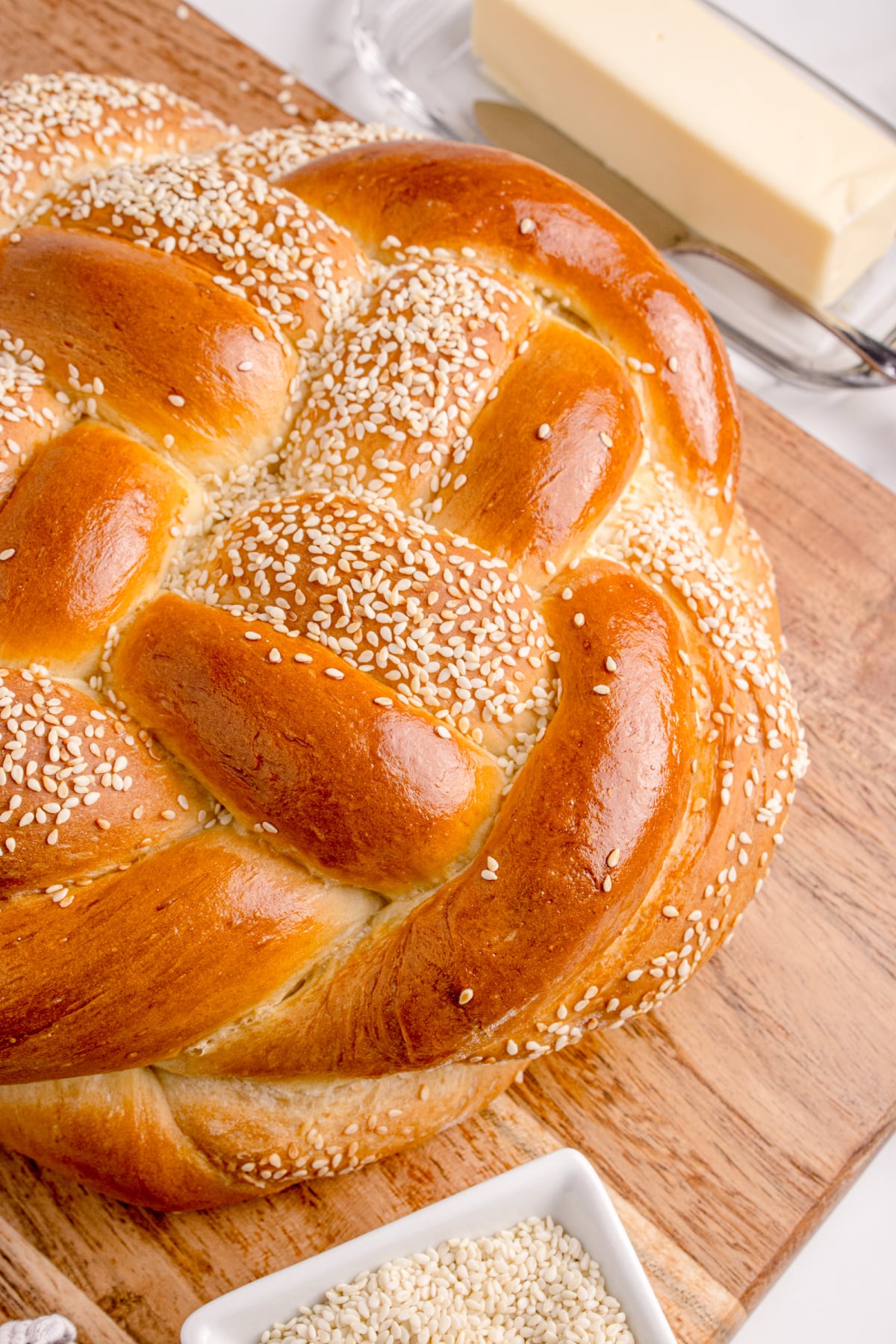 Golden sesame round challah with shiny braided crust Round challah bread on a wooden board with butter and sesame seeds on the side