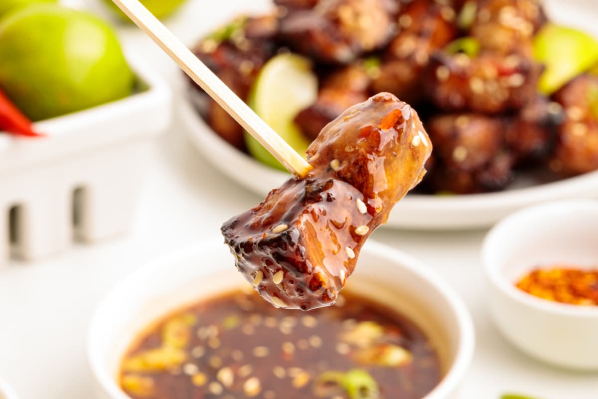 Glazed pork belly bite on a skewer being dipped into sweet chili lime sauce, with the full plate of pork belly in the background.