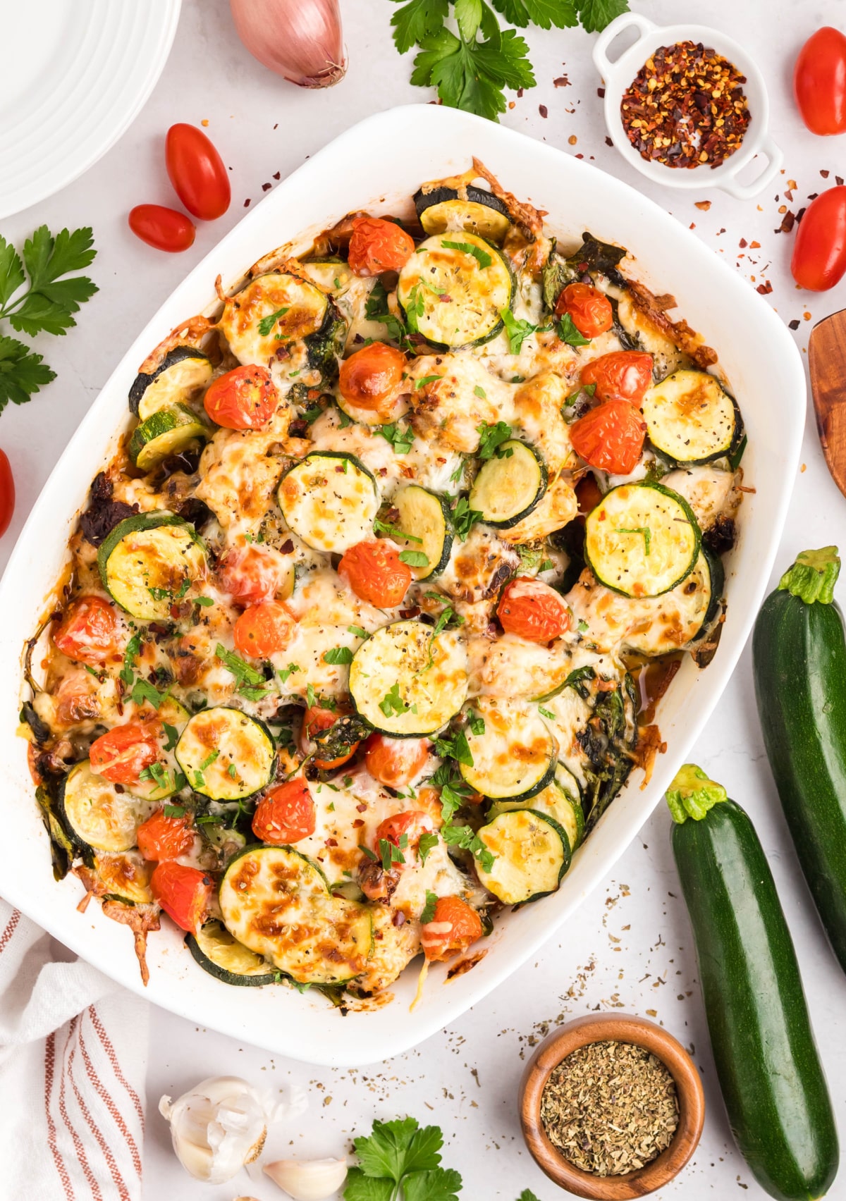 Baked Tuscan Chicken Zucchini Casserole with Mozzarella and Vegetables Overhead view of Tuscan chicken zucchini casserole in white baking dish with golden cheese, tomatoes, and sliced zucchini.