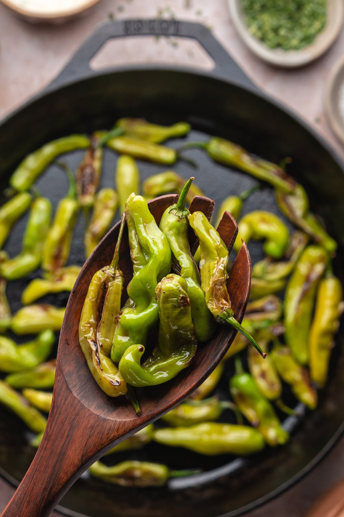 Cast iron blistered shishito peppers in a hot skillet Close-up of blistered peppers on a wooden spoon held over a cast iron pan
