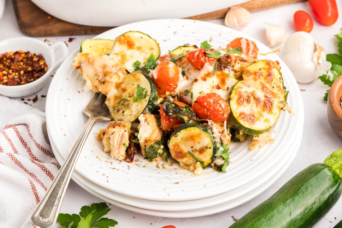 Plated Serving of Cheesy Chicken Zucchini Casserole Cheesy zucchini chicken casserole served on a white plate with tomatoes, spinach, and herbs.