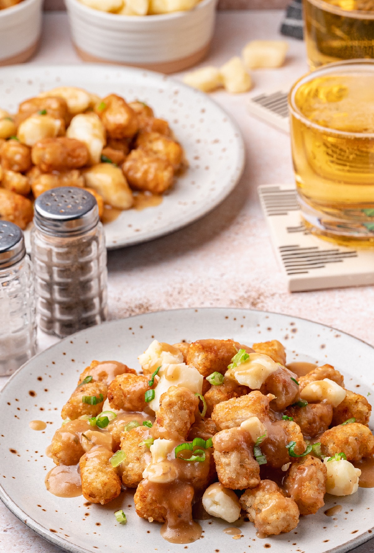 Tater Tot Poutine with Cheese Curds and Homemade Gravy Tater tot poutine served on two plates with cheese curds and gravy, with drinks and salt shakers in the background.