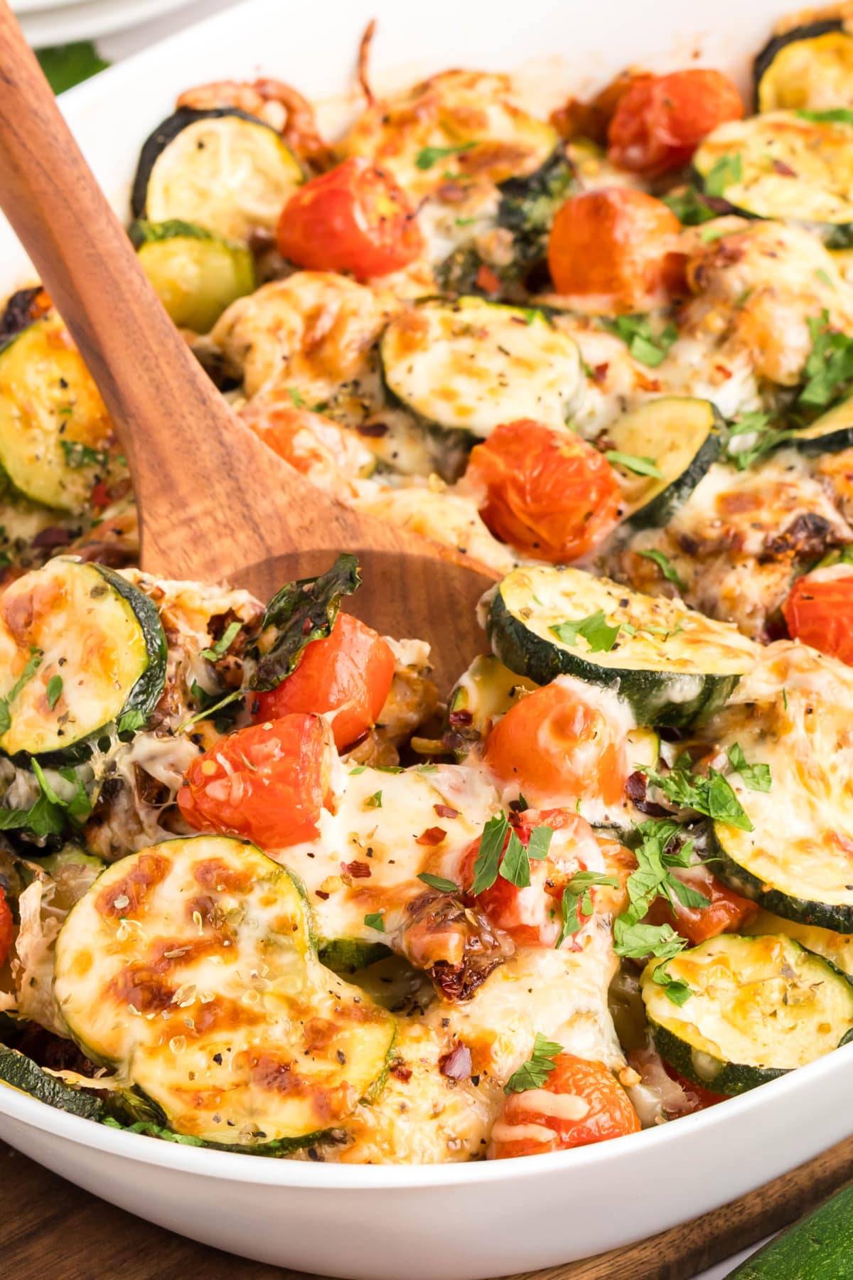One Pan Chicken Zucchini Casserole with Mozzarella and Tomatoes Wooden spoon lifting cheesy chicken and zucchini from casserole dish with roasted tomatoes and herbs.