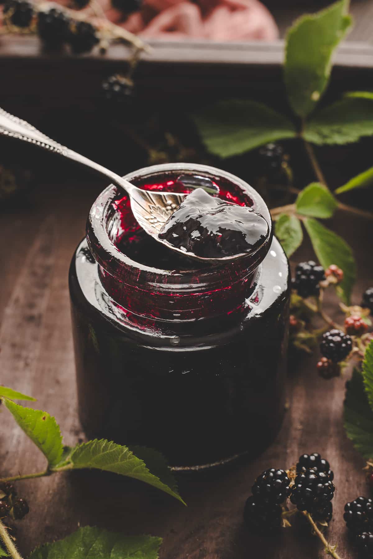 Spoon lifting glossy blackberry jam from a jar surrounded by fresh blackberry leaves