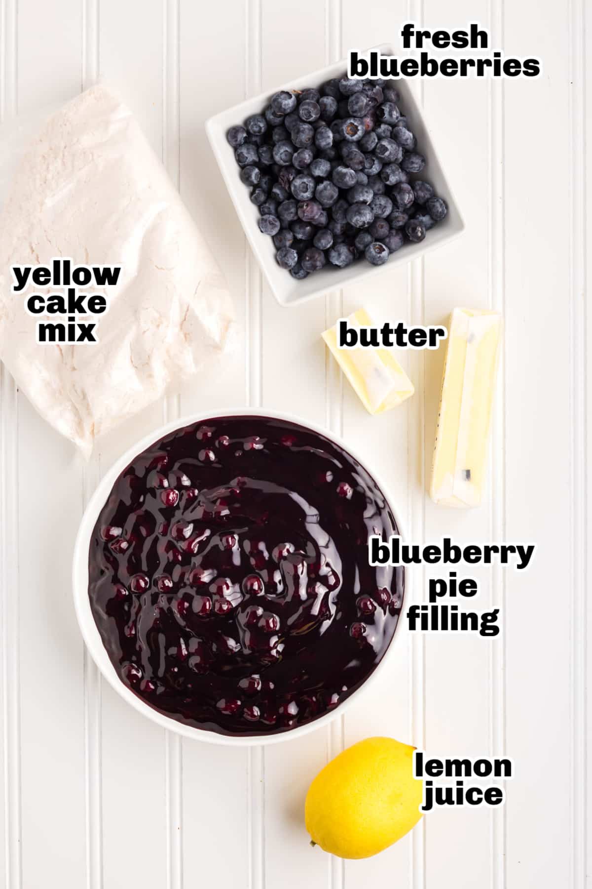 Overhead view of ingredients like cake mix, pie filling, blueberries, lemon, and butter on a countertop.