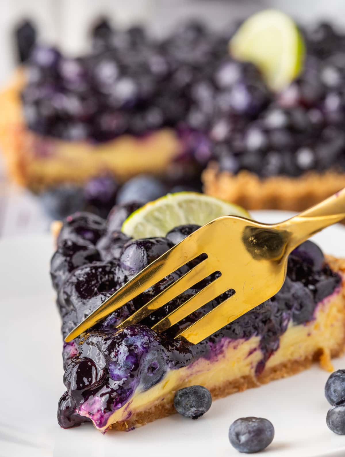 Blueberry Key Lime Tart slice with fork. Bite taken from Blueberry Key Lime Tart slice topped with blueberries and lime.