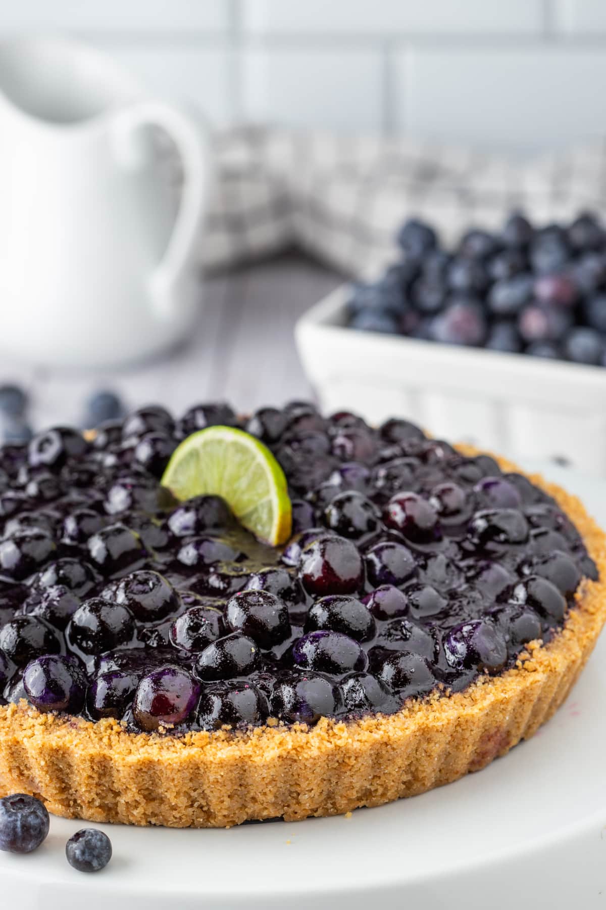 Blueberry lime tart whole on platter. Blueberry lime tart in graham cracker crust topped with fresh blueberries and lime.