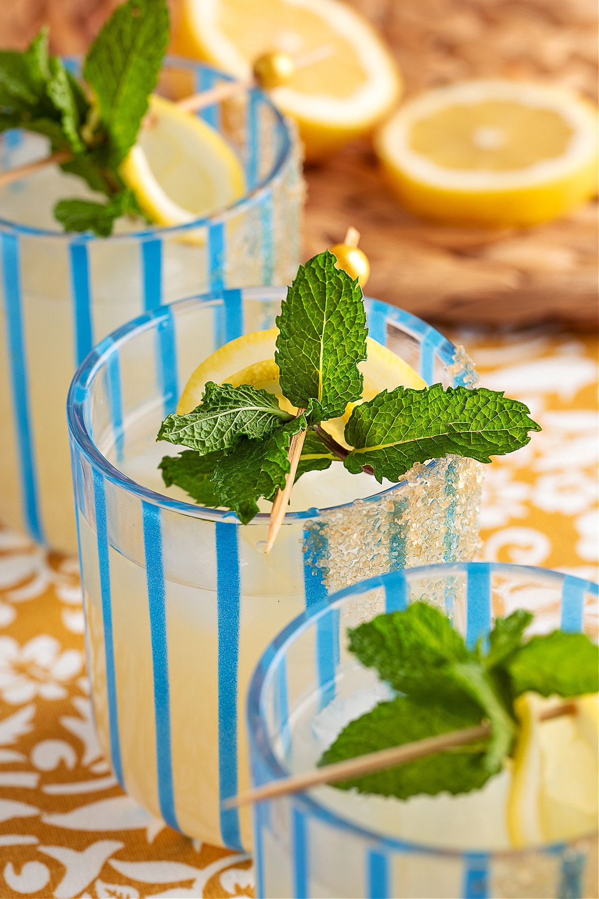Three lemon mint gin cocktails lined up on a patterned tablecloth with sliced lemons in the background.