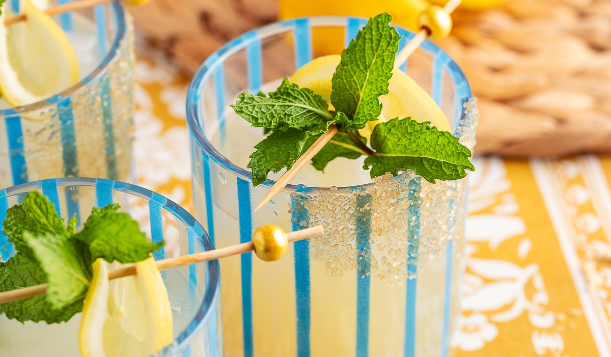 Blue-striped glasses filled with ice and elderflower gin smash, garnished with lemon wedges and fresh mint.