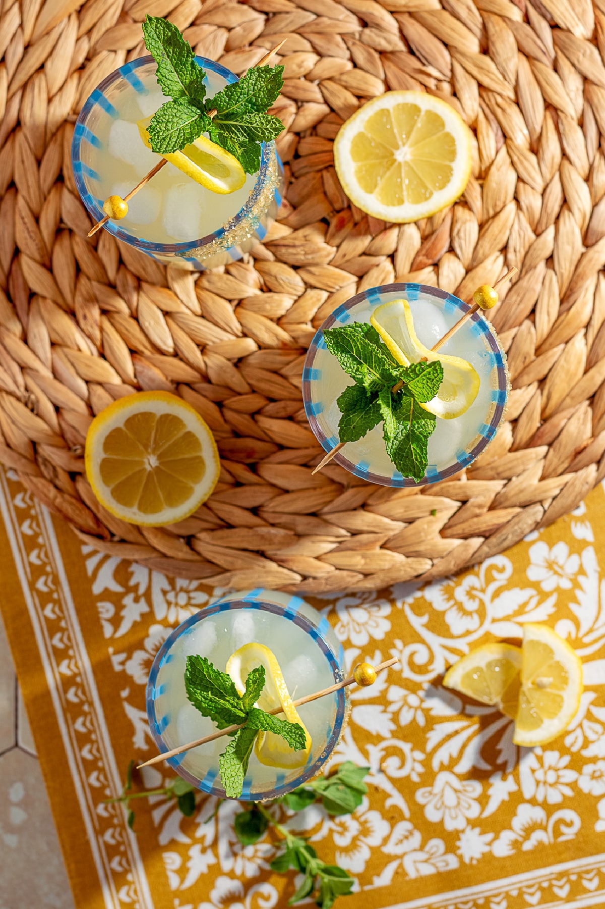 Overhead shot of three honey gin smashes with lemon wheels and mint, arranged on a woven placemat.