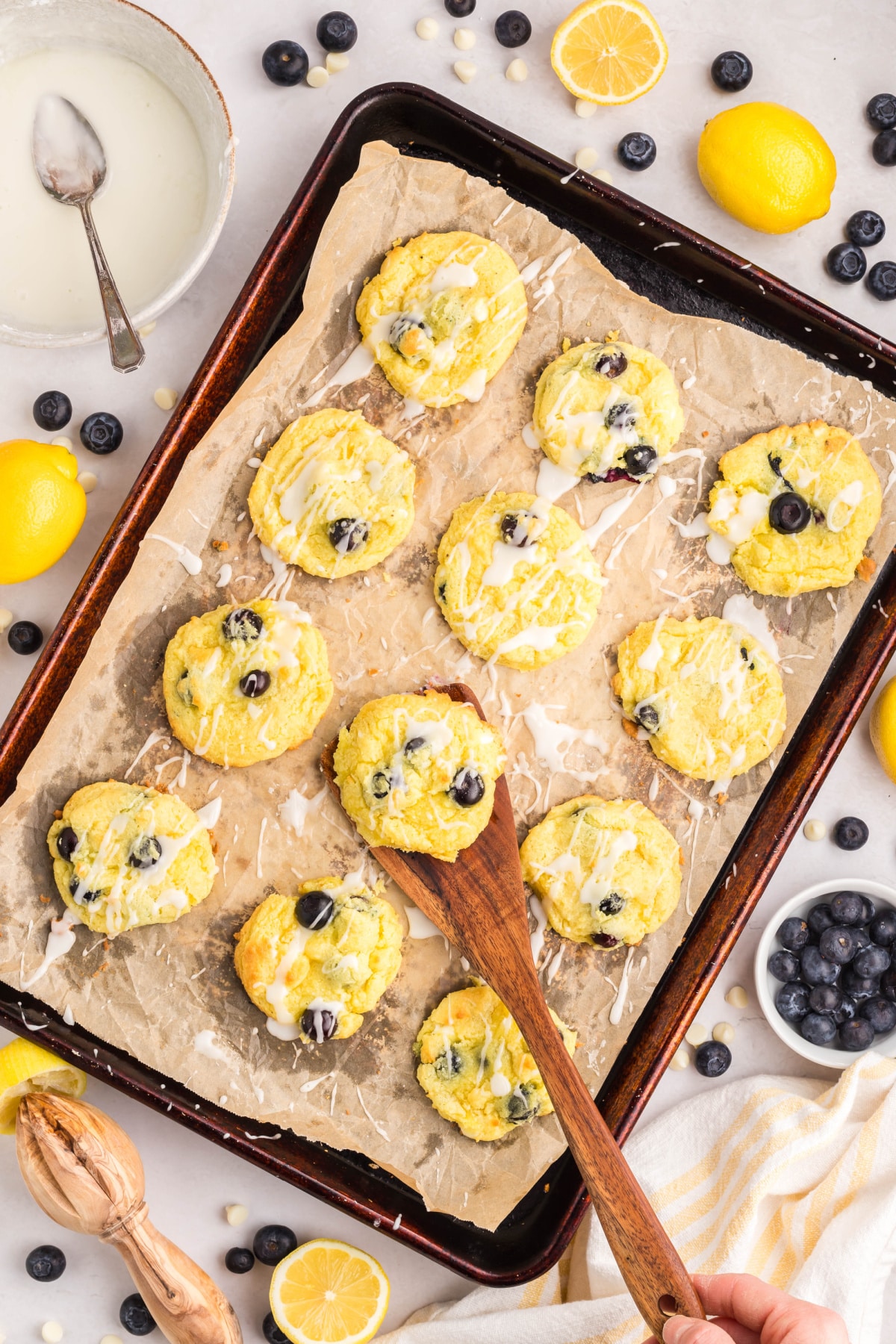 Blueberry Lemon Cake Cookies on Sheet Pan Freshly baked blueberry lemon cookies on parchment with glaze, lemons, and blueberries