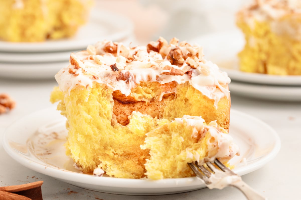 butternut-squash-cinnamon-rolls-bite Butternut squash cinnamon roll on a white plate with cream cheese glaze and pecans, a fork pulling away a bite.