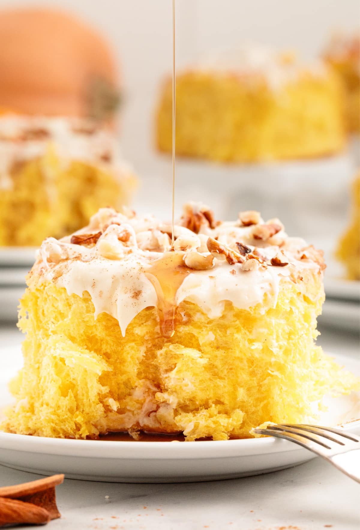 butternut squash cinnamon rolls with maple drizzle Close-up of a butternut squash cinnamon roll topped with cream cheese glaze, chopped nuts, and maple syrup being drizzled on top.