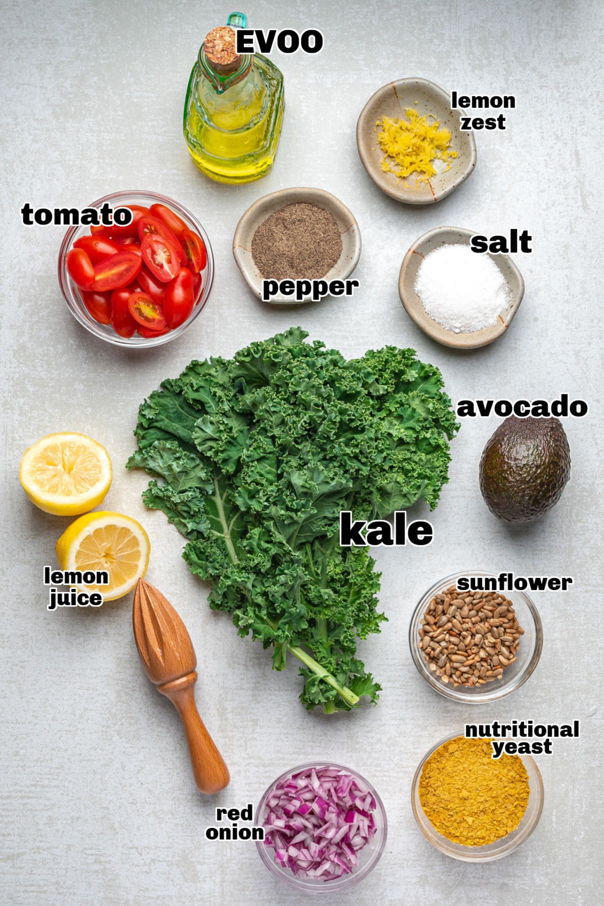 Ingredients for avocado kale salad with “cheesy” flavor arranged on a surface with kale, avocado, lemon, tomatoes, sunflower seeds, and nutritional yeast.