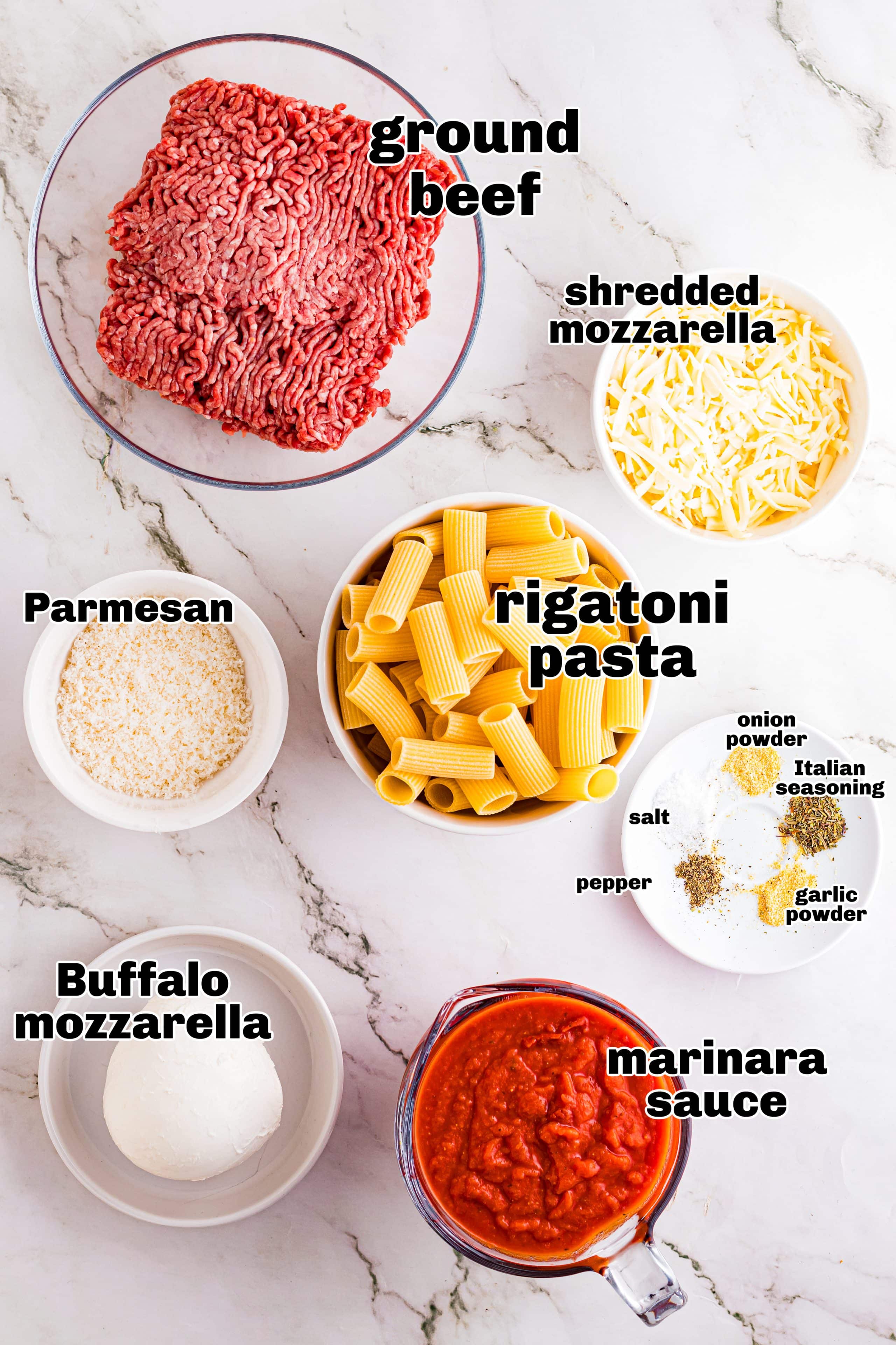 Ingredients for baked rigatoni including ground beef, rigatoni pasta, mozzarella, Parmesan, marinara sauce, and seasonings arranged on a marble surface