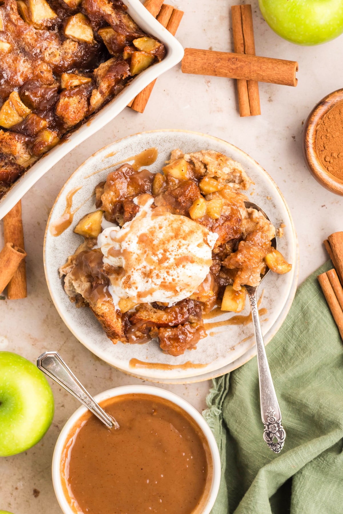 Overhead view of baked brioche apple dessert topped with ice cream and caramel sauce surrounded by cinnamon sticks and green apples.