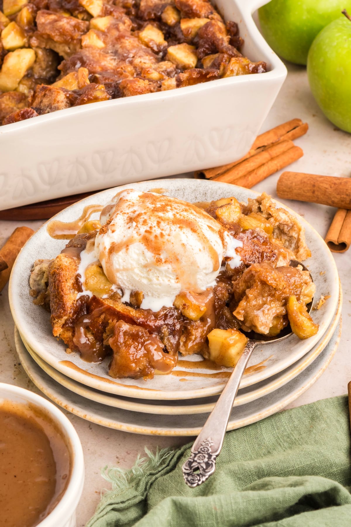 Warm brioche apple dessert topped with vanilla ice cream and rich caramel sauce, surrounded by cinnamon sticks and green apples.