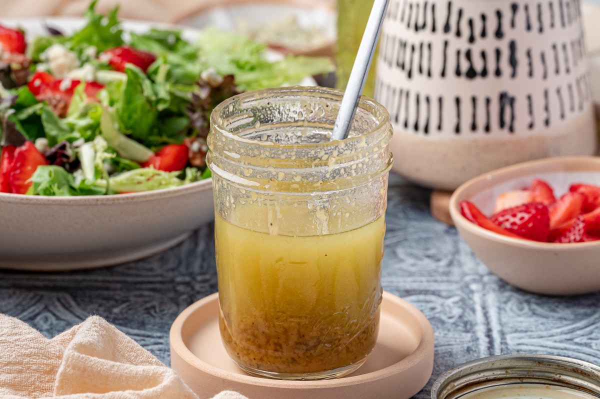Homemade Champagne Vinaigrette with Salad Greens Homemade champagne vinaigrette in a jar with spoon, surrounded by fresh salad greens and sliced strawberries.