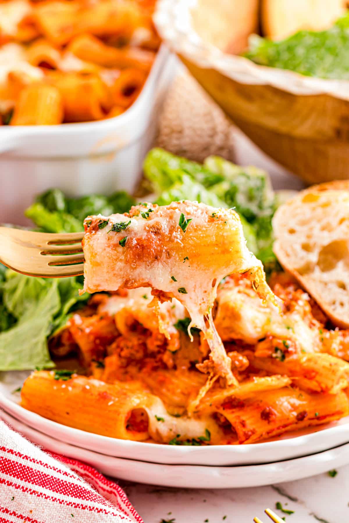 Forkful of cheesy rigatoni with meat sauce and melted mozzarella served with salad and bread on a plate