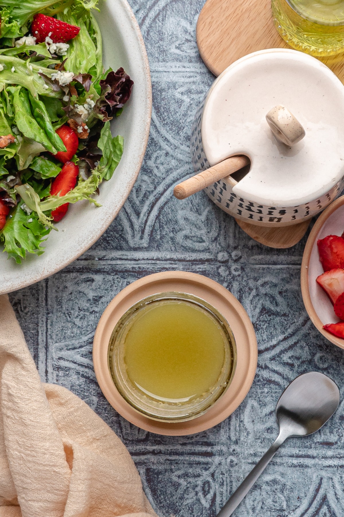 Dijon Champagne Dressing with Fresh Salad Glass jar of Dijon champagne dressing next to a strawberry and mixed greens salad on a textured blue surface.