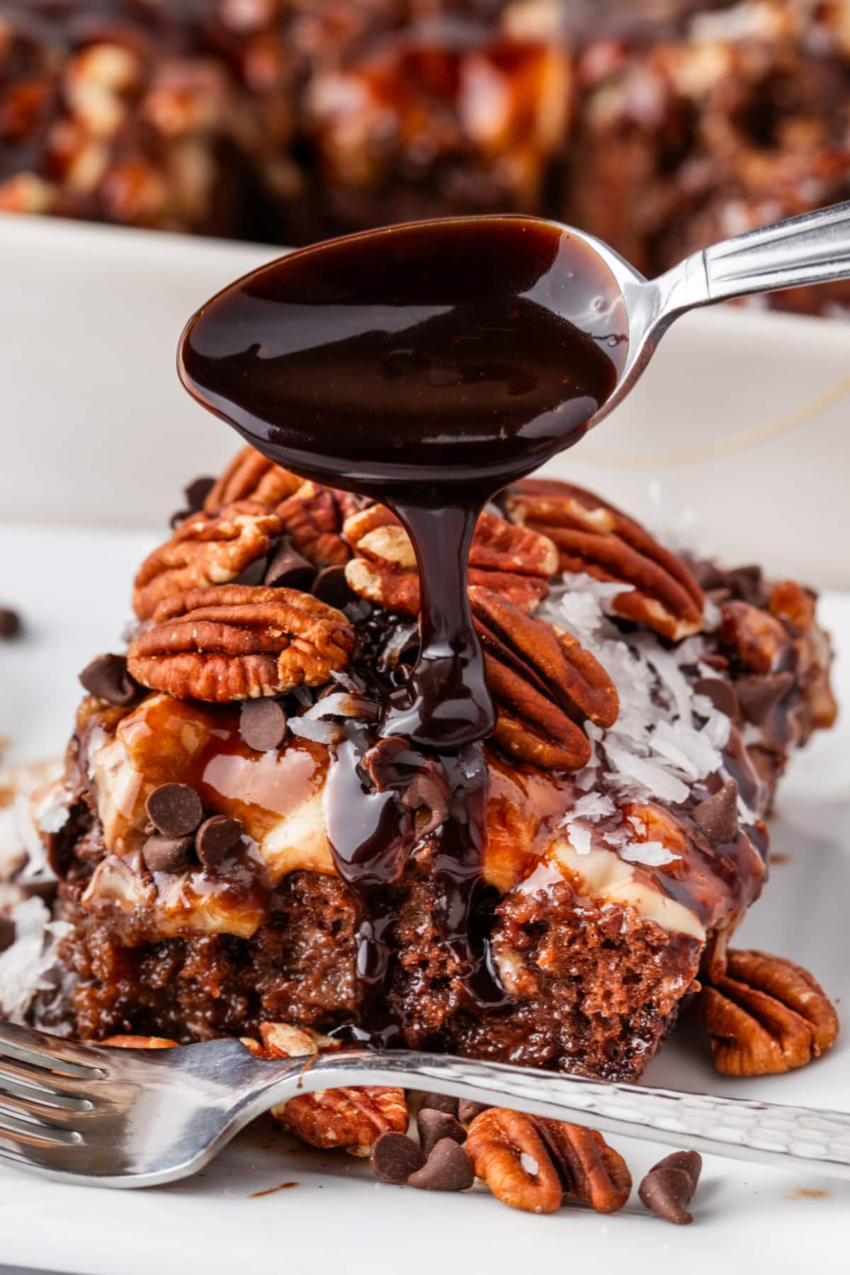 German chocolate poke cake with chocolate drizzle, pecans, and coconut German chocolate poke cake slice topped with pecans and coconut as warm chocolate sauce drips from a spoon over the top.