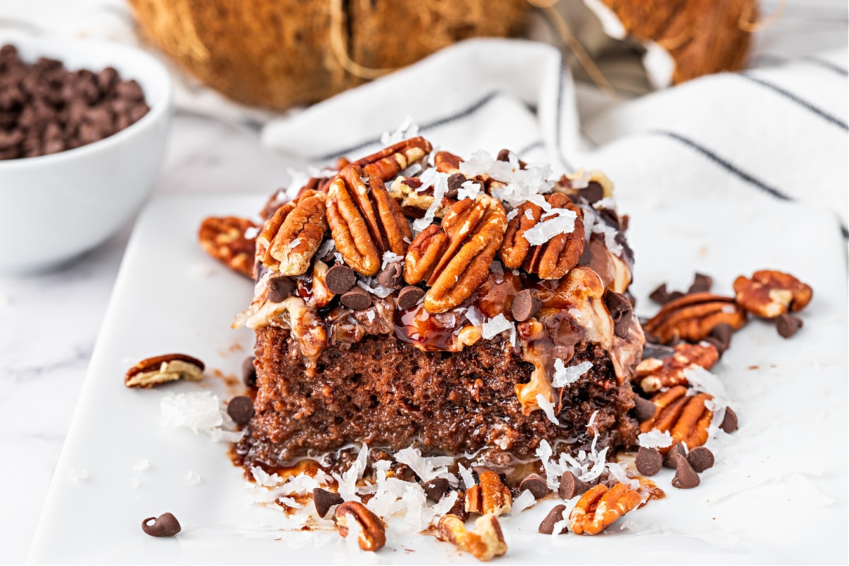 German chocolate poke cake slice with caramel and pecans Single serving of German chocolate poke cake on plate with caramel drizzle, coconut, pecans, and chocolate chips.