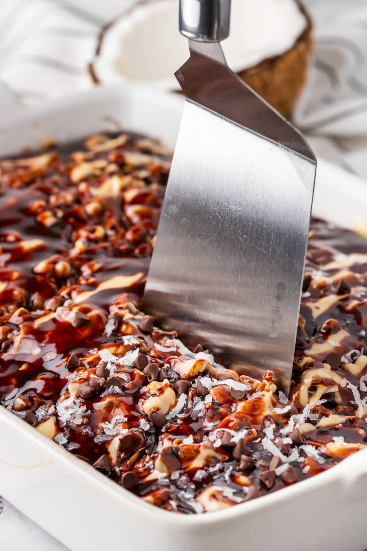 German chocolate poke cake in pan with caramel, coconut, and pecans German chocolate poke cake in a baking pan topped with caramel drizzle, shredded coconut, pecans, and chocolate chips with a spatula ready to serve.