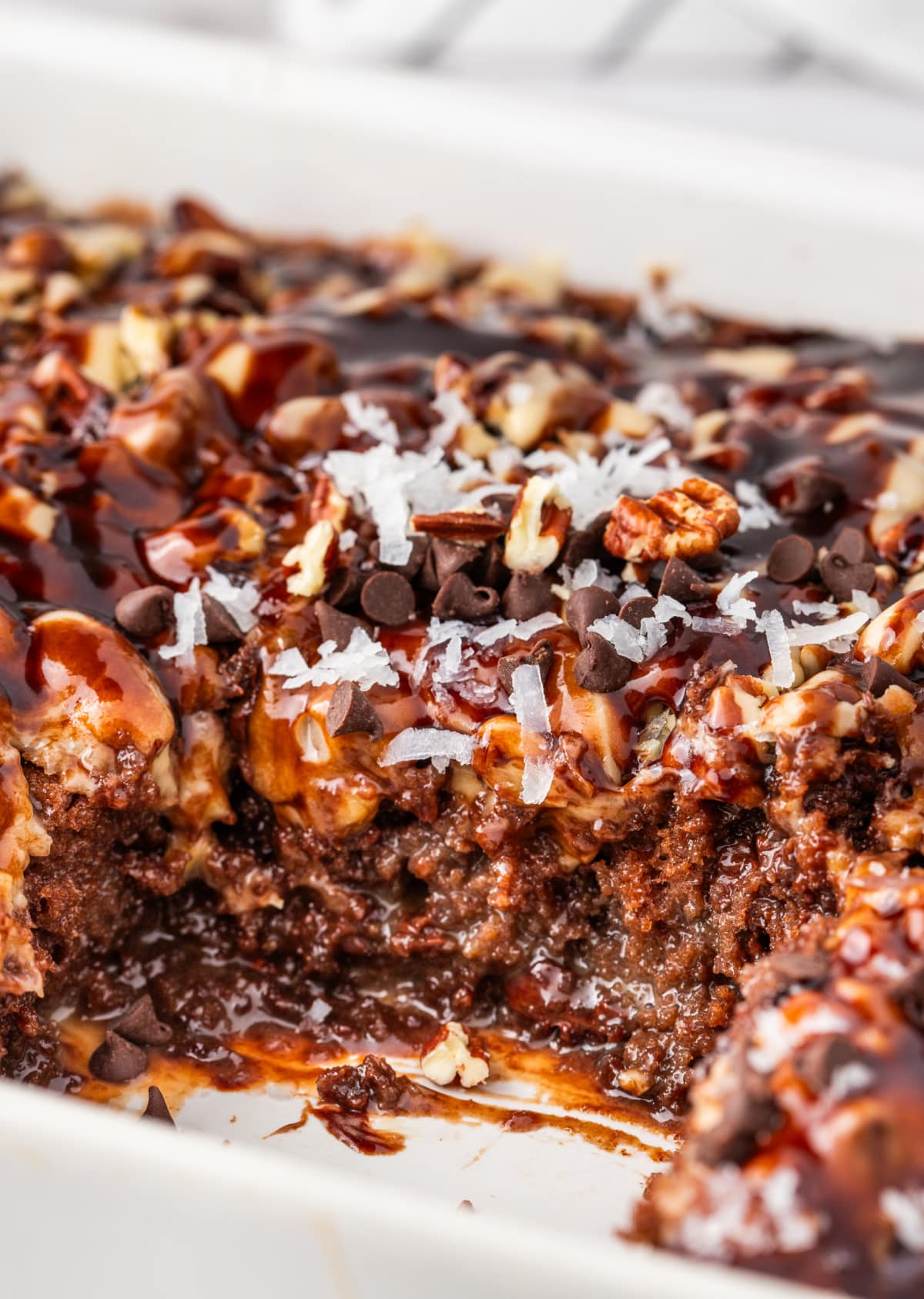 Chocolate caramel poke cake in pan with coconut, pecans, and chocolate chips Close-up of chocolate caramel poke cake in a pan showing gooey layers of caramel, shredded coconut, pecans, and melted chocolate chips.