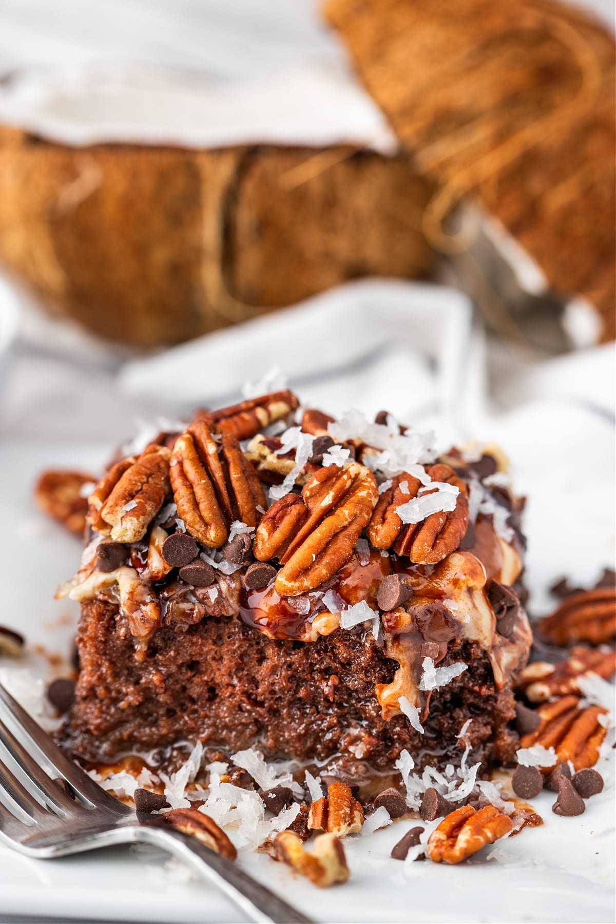 German chocolate poke cake slice with pecans and coconut German chocolate poke cake slice on a white plate topped with pecans, shredded coconut, and chocolate chips.