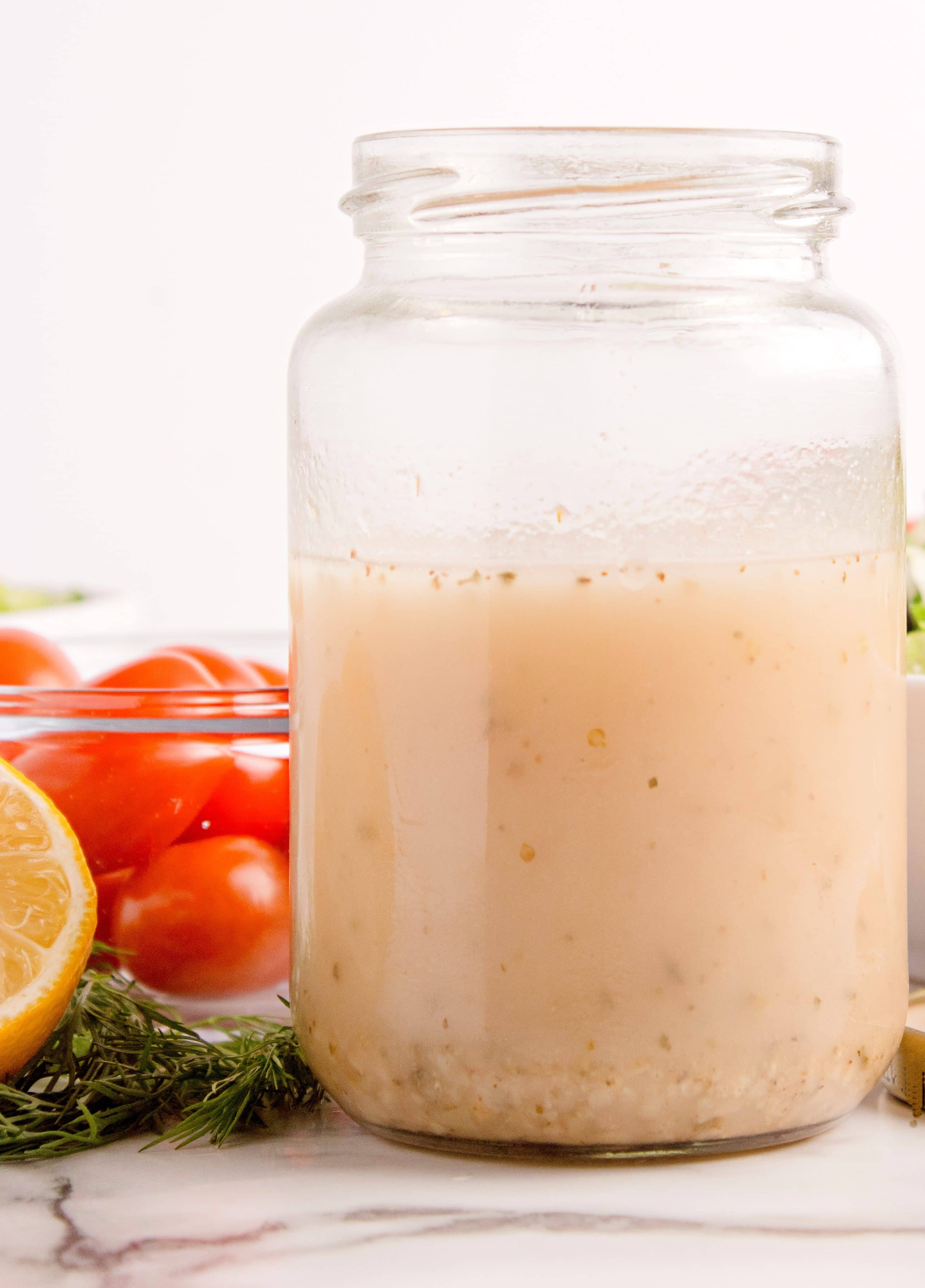 Side view of a glass jar filled with creamy Greek salad dressing and tomatoes and lemon in the background.