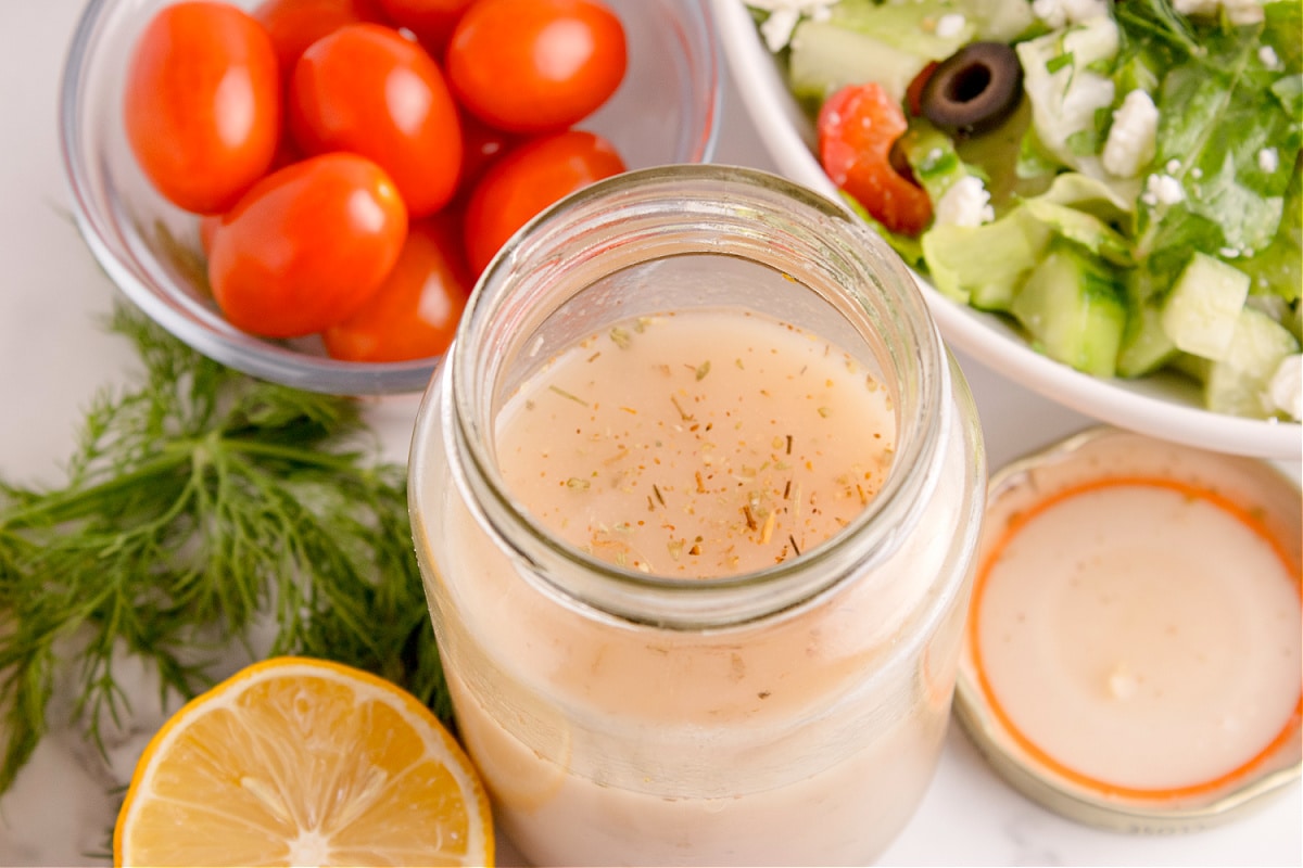 Greek salad dressing in a jar beside a bowl of chopped Greek salad with tomatoes, olives, and feta cheese.