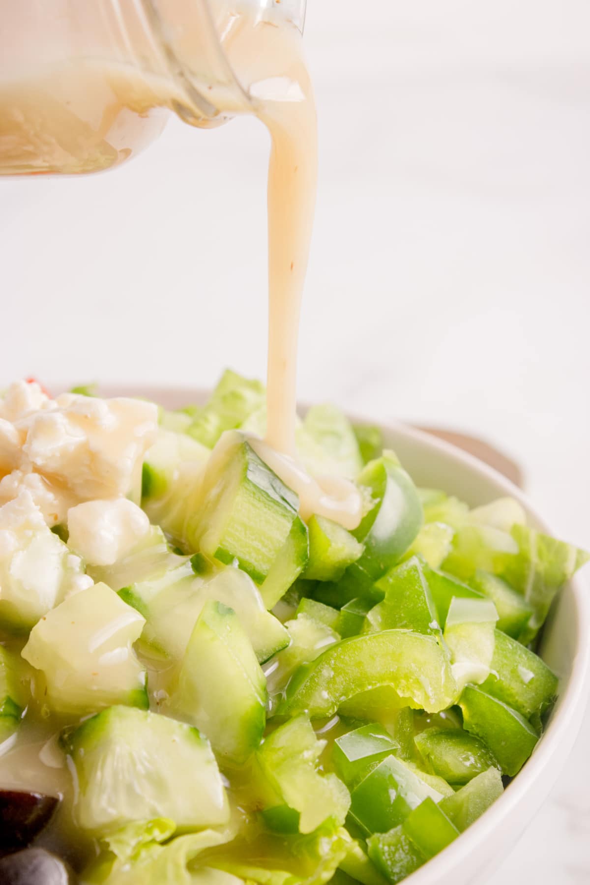 Greek salad dressing being poured from a jar over a bowl of chopped cucumbers, peppers, and feta.