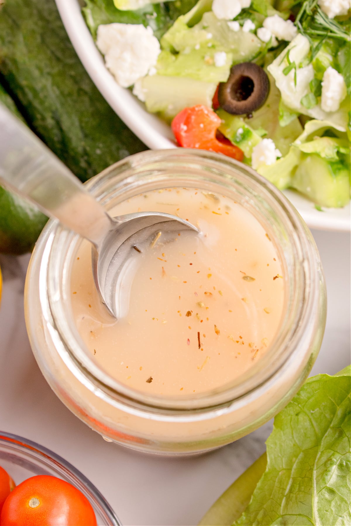 Jar of homemade Greek vinaigrette with a spoon beside a Greek salad topped with feta, olives, and tomatoes.