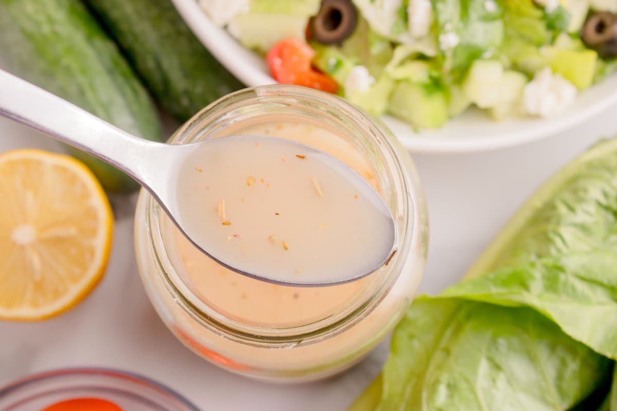 Spoon lifting lemon Dijon Greek dressing from a glass jar, surrounded by fresh salad ingredients.