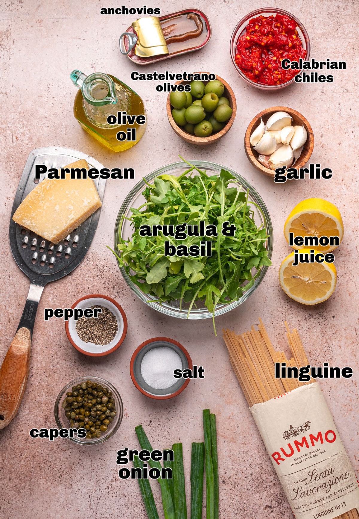 green-olive-puttanesca-pasta-ingredients-castelvetrano-anchovy-chilies Ingredients for green olive puttanesca pasta arranged on a pink surface with Castelvetrano olives, anchovies, Calabrian chilies, arugula, lemon, and linguine.