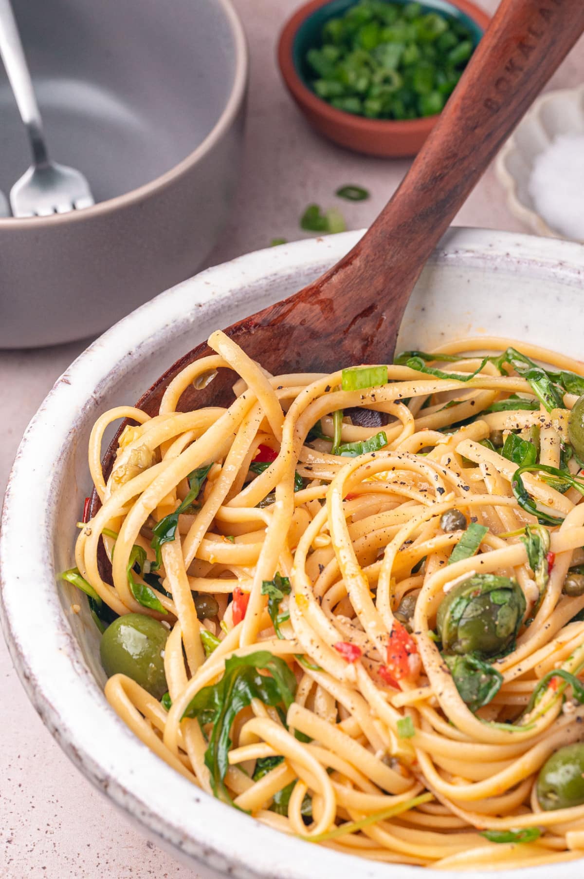 olive-and-caper-pasta-with-arugula-and-capers Olive and caper pasta in a white serving bowl topped with arugula, green onions, and Castelvetrano olives.