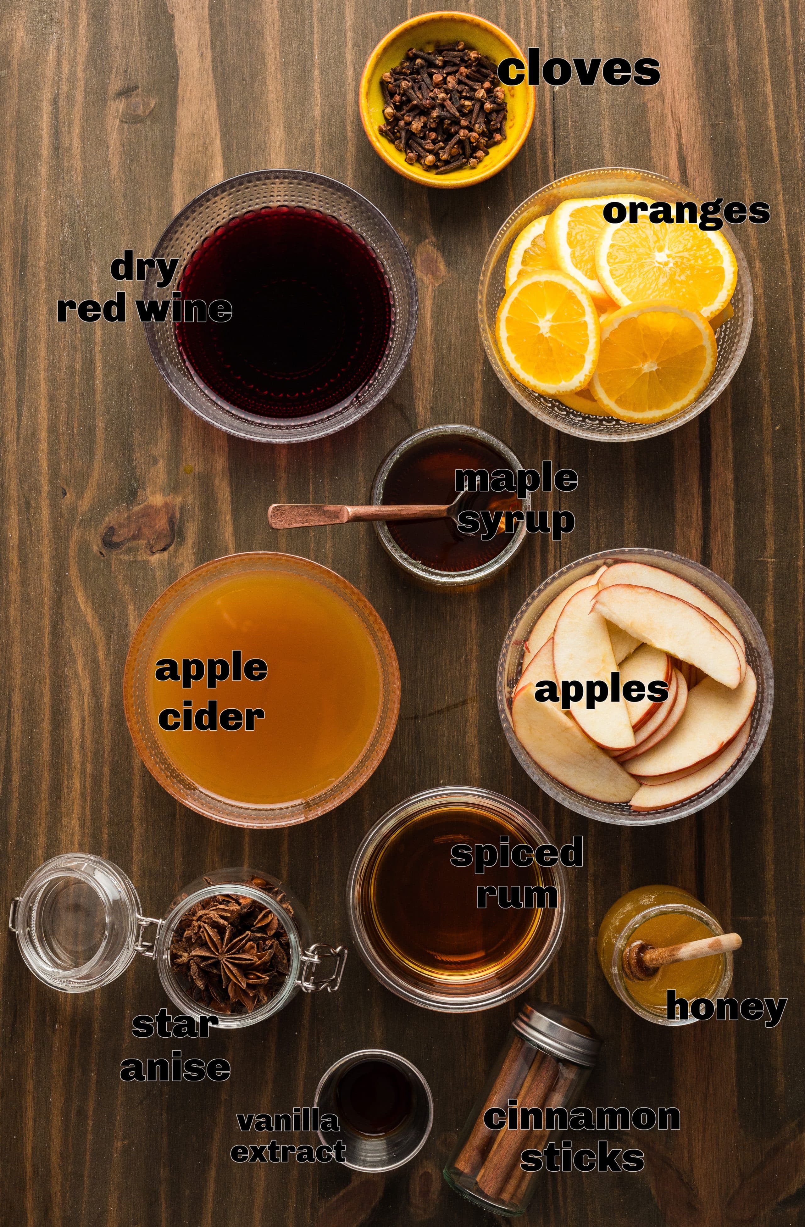 Ingredients for mulled apple cider arranged on a wooden table including apple cider, red wine, rum, maple syrup, honey, oranges, apples, cinnamon sticks, cloves, star anise, and vanilla extract.