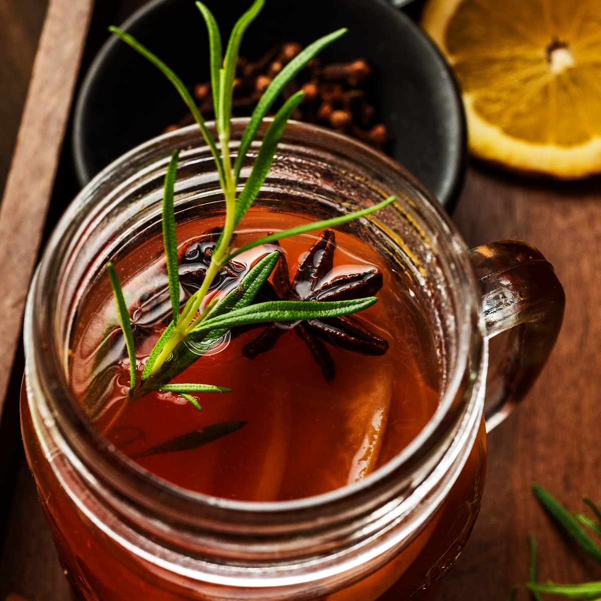 Glass mug of mulled cider garnished with rosemary and star anise beside sliced lemon and cloves, photographed for Noble Pig.