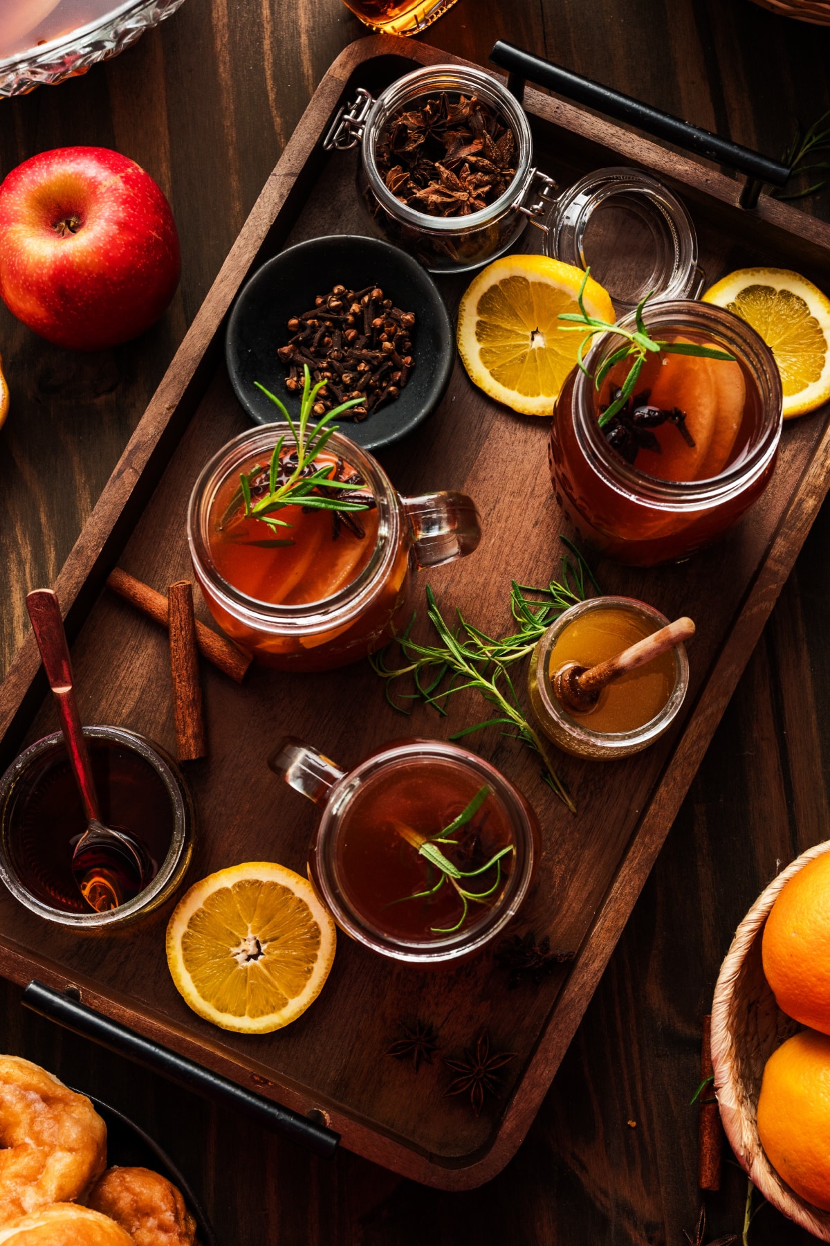Tray of warm cider mugs surrounded by spiced rum, oranges, honey, cinnamon sticks, rosemary, and star anise, highlighting the ingredients that flavor the mulled cider.