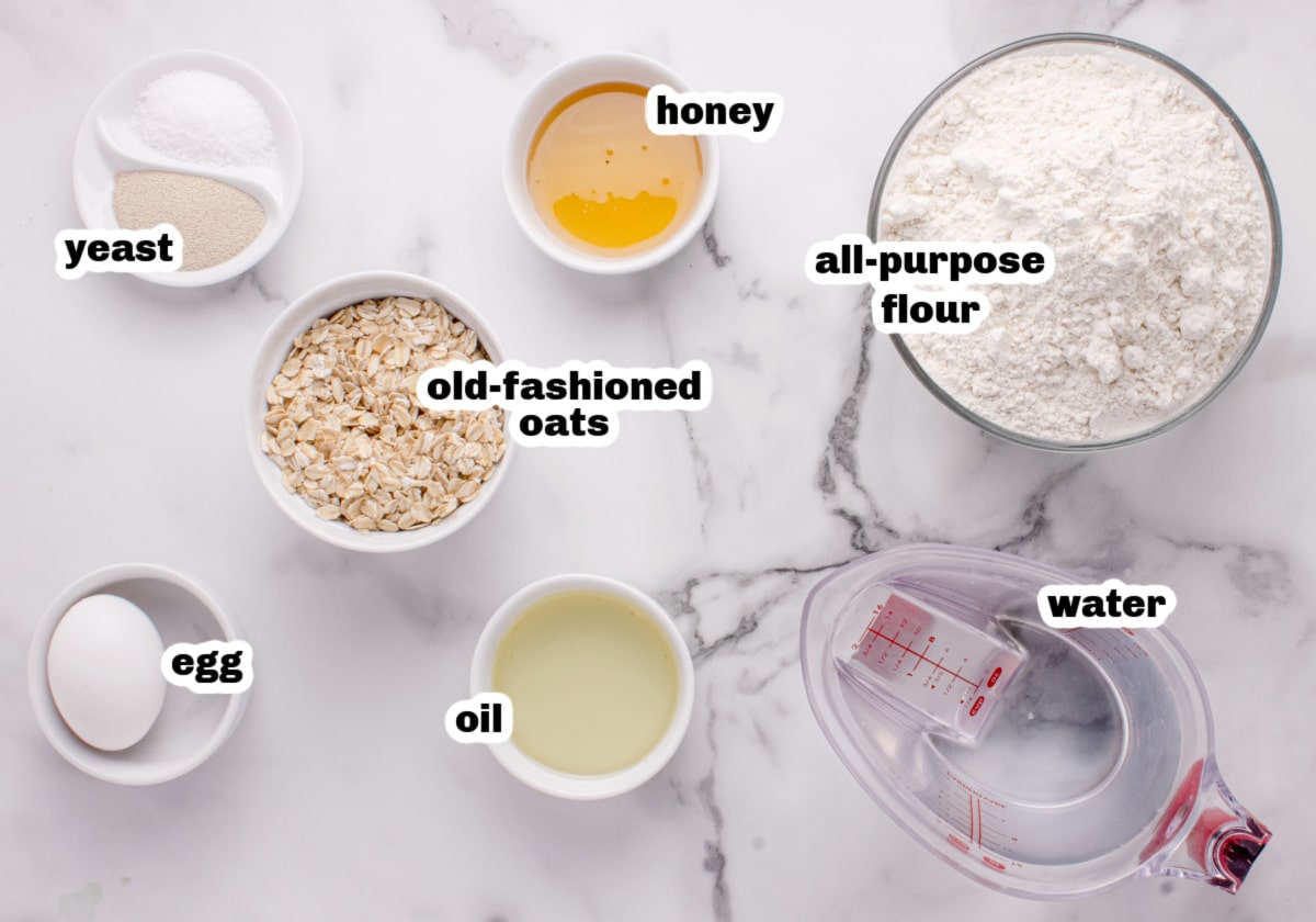 honey oatmeal bread ingredients Ingredients for homemade oatmeal bread including oats, flour, honey, yeast, oil, and egg on a marble counter.