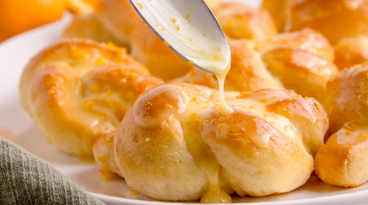 Spoon drizzling orange glaze over freshly baked rolls.
