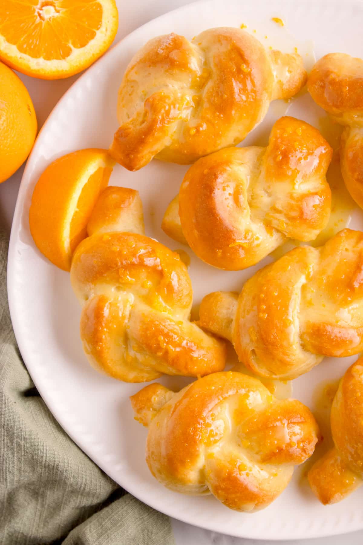 Plate of golden orange knot rolls with shiny glaze and sliced oranges beside them.
