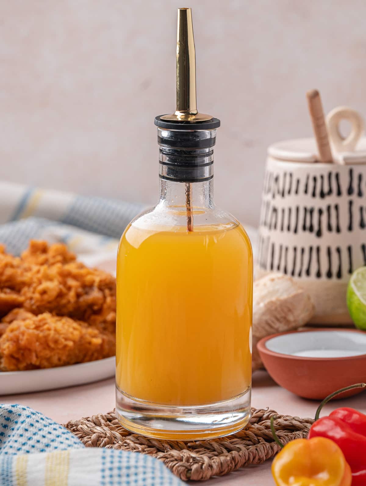 Clear glass bottle filled with golden homemade hot sauce, with fried chicken and habanero peppers in the background.