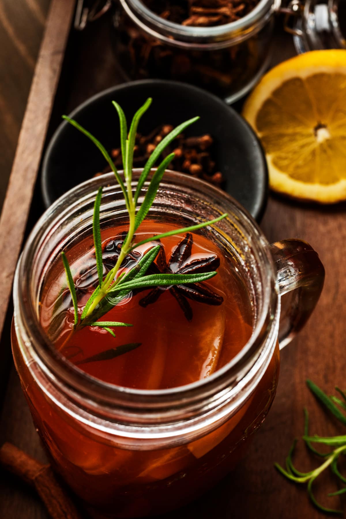 Glass mug of mulled apple cider topped with rosemary and star anise beside lemon slices and cloves, showing the drink’s deep color and aromatic garnish.