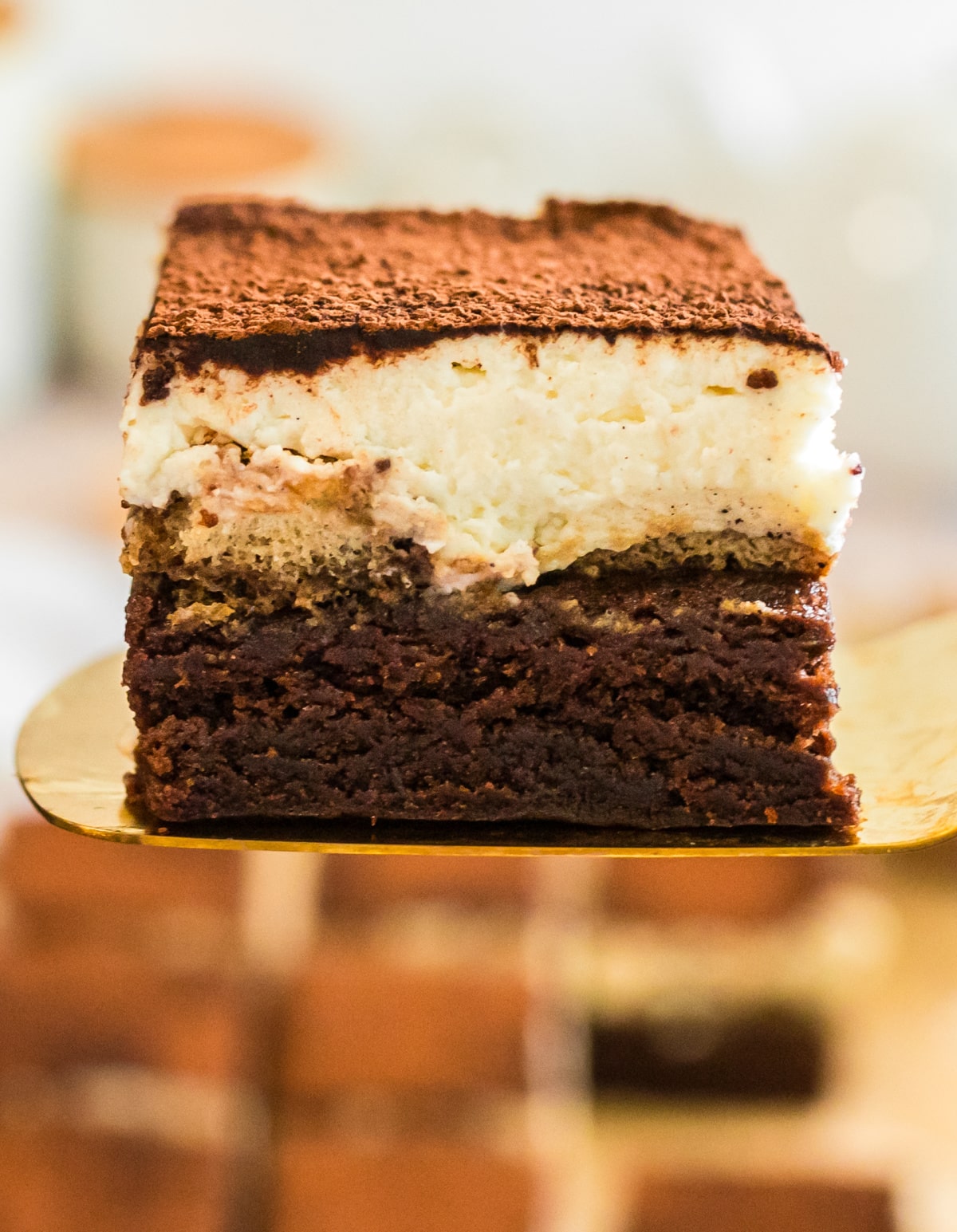 slice of tiramisu brownies on a gold spatula with distinct brownie, ladyfinger, and mascarpone layers.