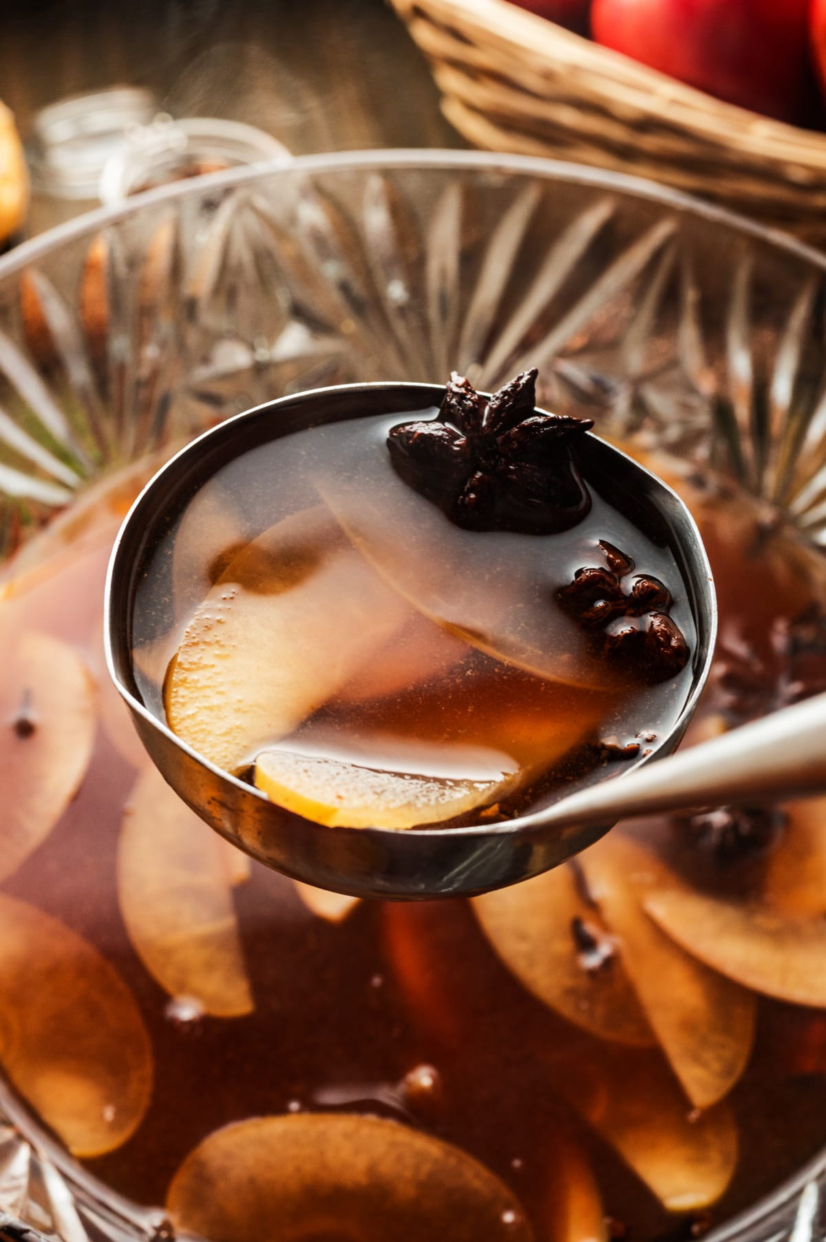 Ladle lifting warm cider with apple slices and star anise from a glass punch bowl, showing the color and spice of the mulled cider.