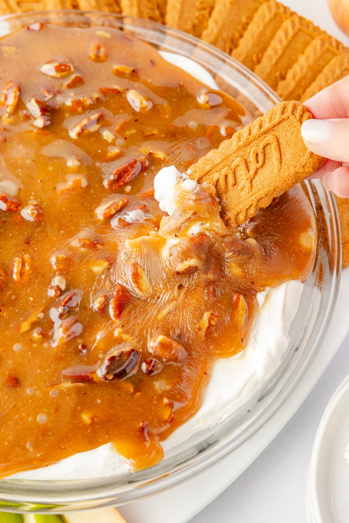 Biscoff cookie dipped into caramel pecan topping over cream cheese layer Pecan pie dip being scooped with a Biscoff cookie, showing the warm caramel pecan topping pulling away from the cold cream cheese layer.