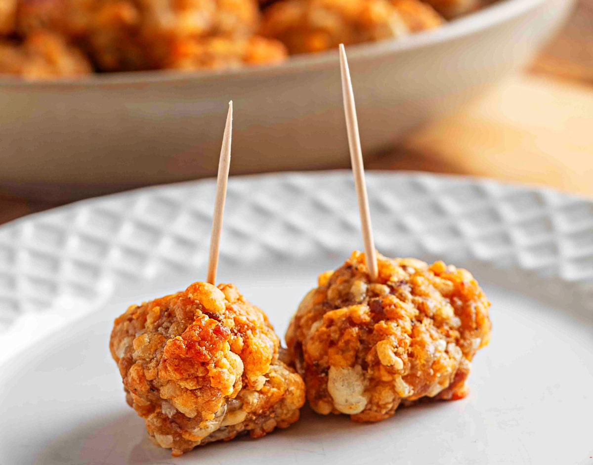 Two sausage balls with toothpicks set on a white plate. Two smoked cheddar sausage balls with toothpicks stand on a white plate, showing their crisp baked edges and tender Bisquick binding.