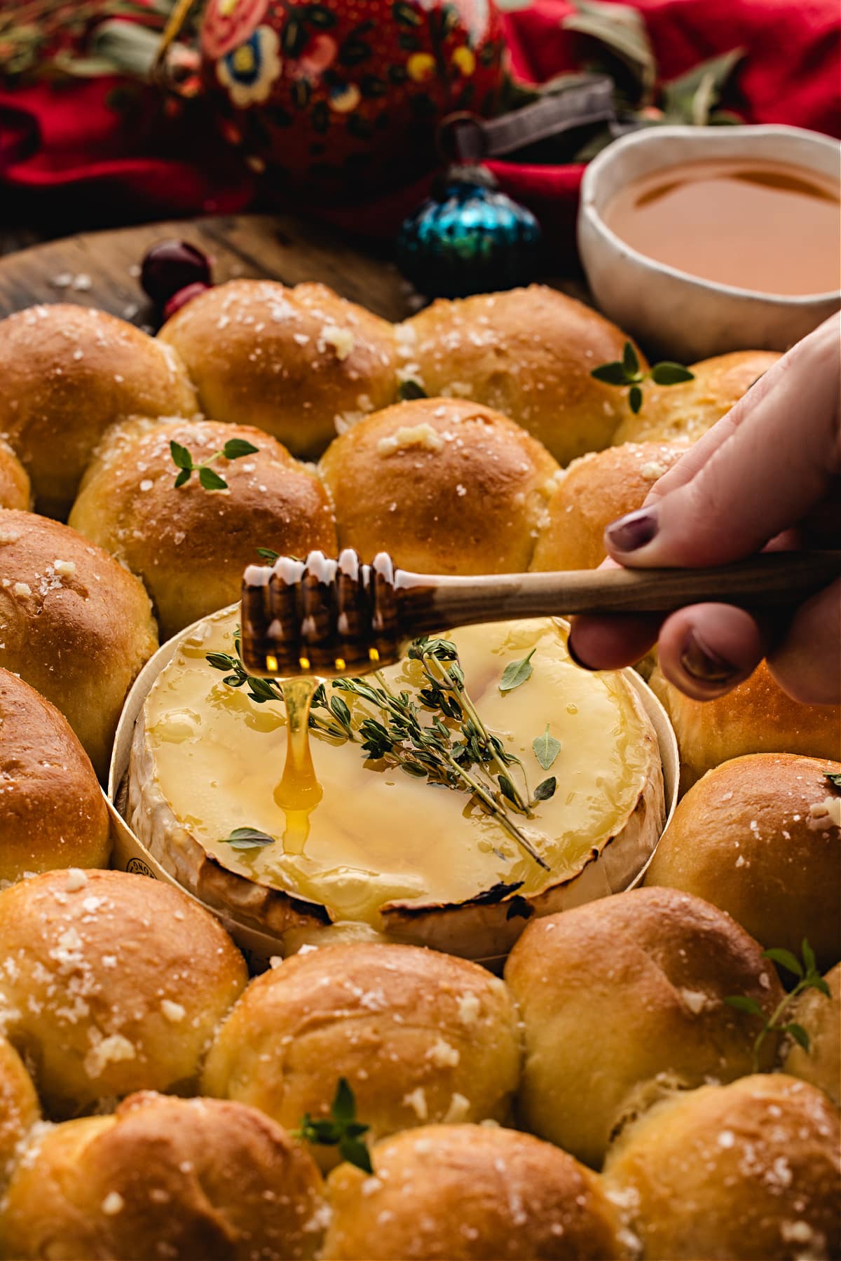 A baked Camembert bread roll wreath with warm golden rolls surrounding a pool of melted cheese, as honey is drizzled over thyme on top.