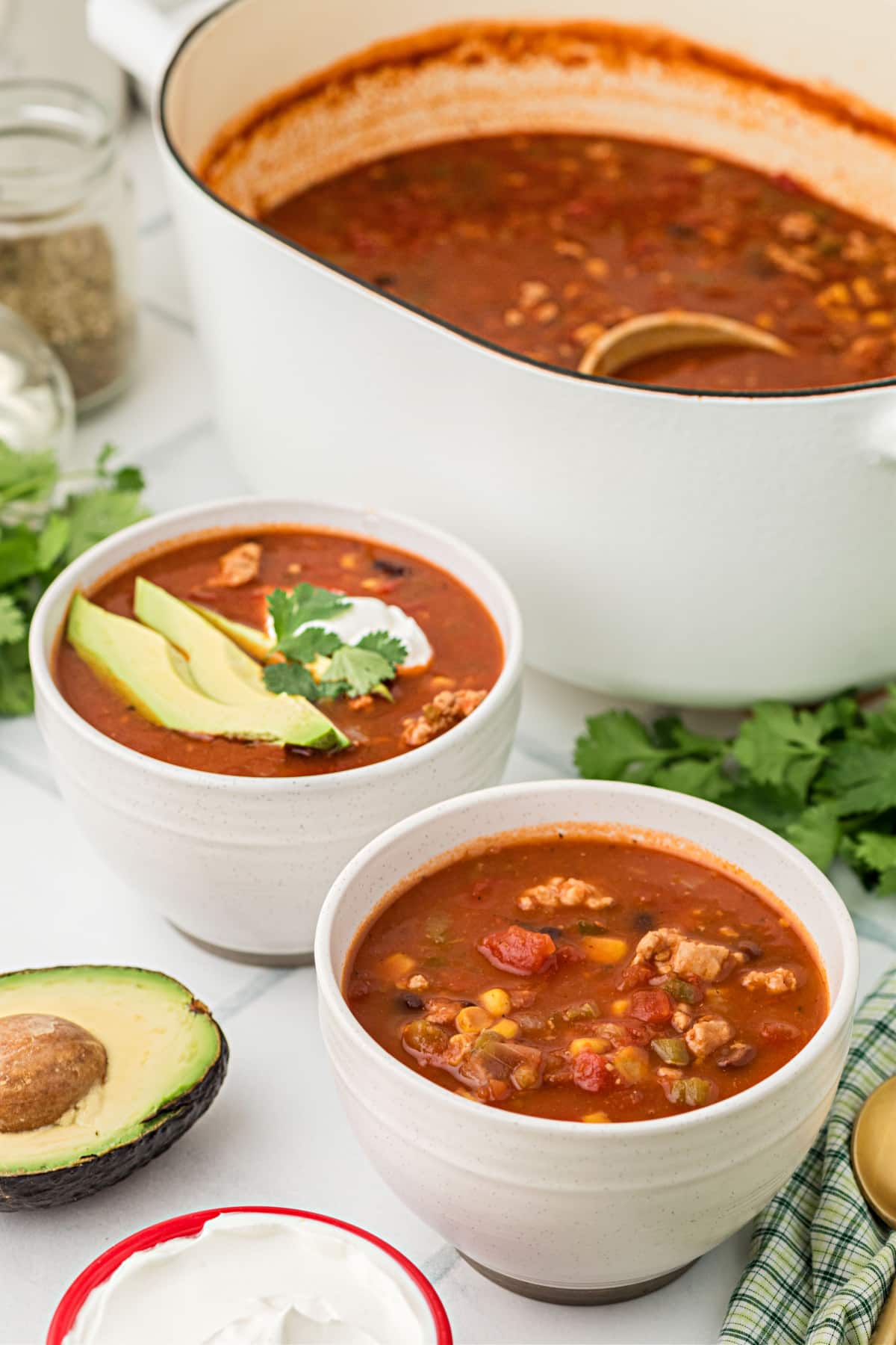 Two bowls of chicken chili topped with avocado, sour cream, and cilantro beside a Dutch oven filled with more chili.